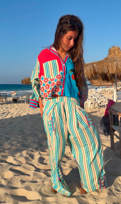 A woman stands on a sandy beach wearing a colorful patchwork-style two-piece outfit with vertical stripes in turquoise, cream, and red, plus a bright turquoise top with red and orange accents. Beach umbrellas, chairs, ocean, and blue sky are visible in the background.